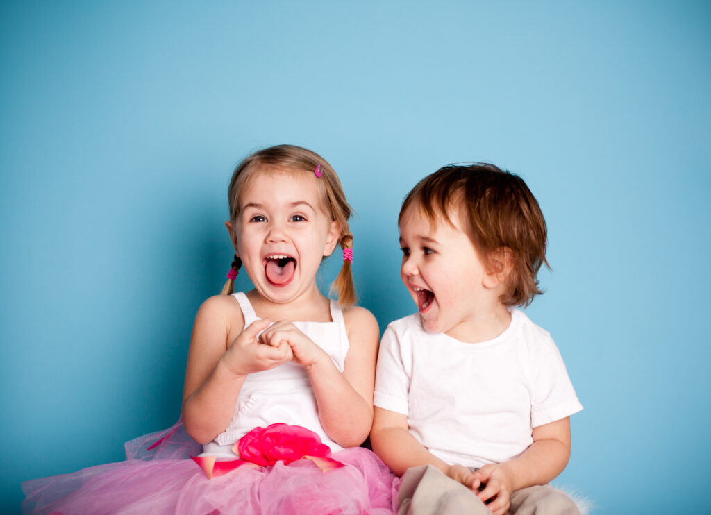 Two young children smiling, looking at each other, blue background, both happy with mouths open in excitement, Kids Smiles Pediatric Dentistry