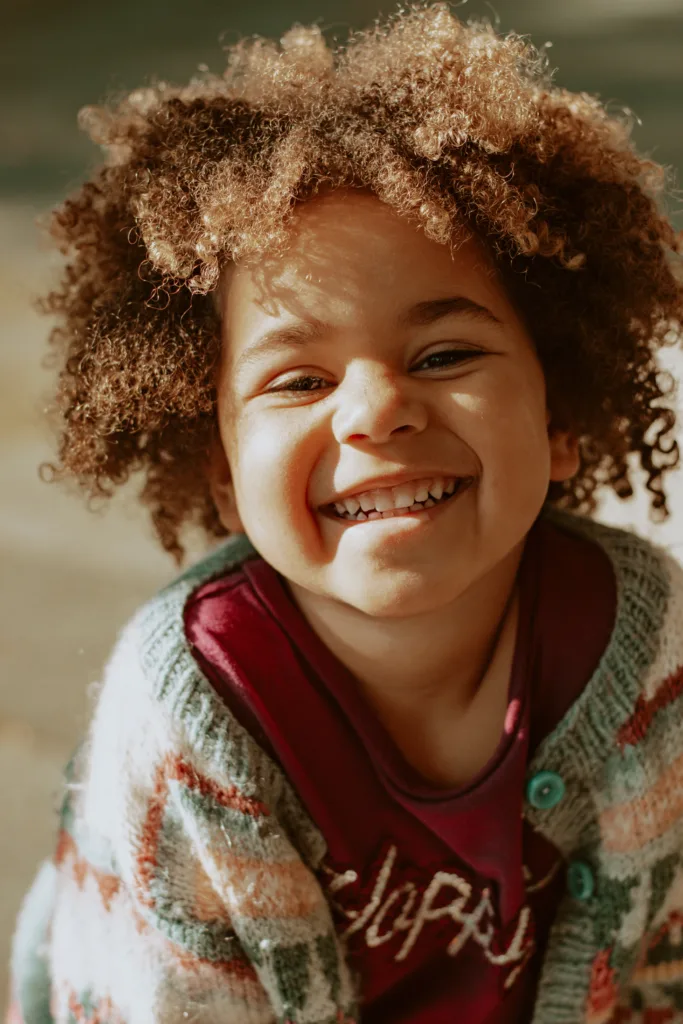Young smiling child with brown curly hair, wearing sweater, cheesing at the camera, Kids Smiles Pediatric Dentistry